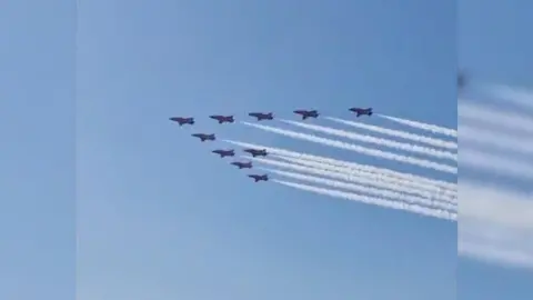 Red Arrows aircraft flies in a ten formation with white smoke trail out the back, blue sky and crowds looking from the ground.