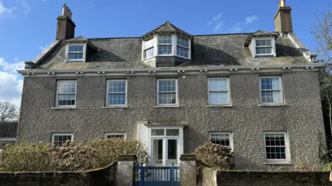 BBC A large rectory in St Martin. The building has a large white door and four windows on the ground floor, five windows on the first floor and three on the third floor. A small white gate leads into the property.
