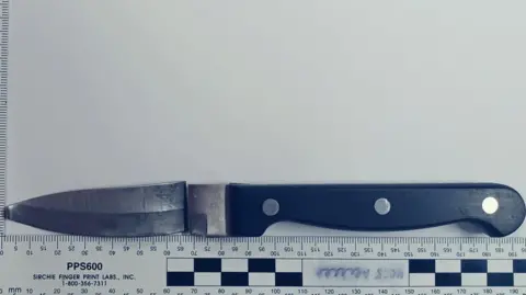 Crown Prosecution Service A black handled kitchen knife, the blade has been snapped away from the hilt but is placed above oit. Below is a ruler measuring the blade and handle to be 195cm long