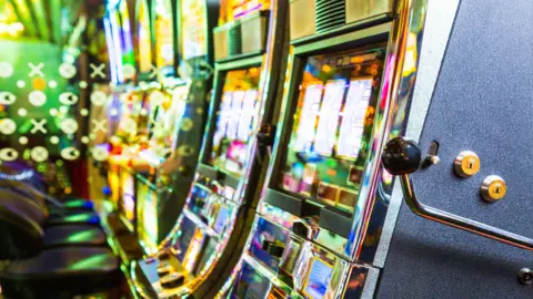A close-up of a row of brightly lit slot machines. The machines are sitting side‑by‑side in a tight line, their reflective chrome edges catching the ambient neon.