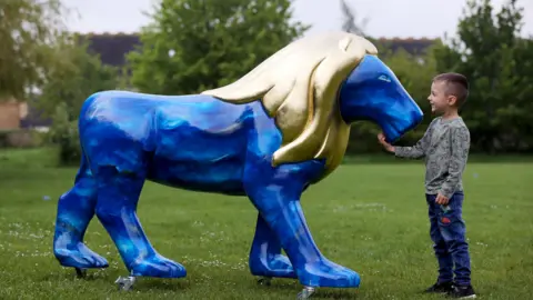 Anna Lythgoe A boy smiles as he touches the chin of a blue lion sculpture with a gold mane in a park
