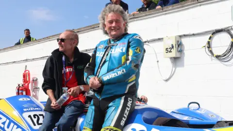 BBC John, who is wearing his leathers, sits on his blue and yellow sidecar, smiling, next to another man. 