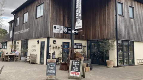 Image of the outside of the Farmers Fayre shop and restaurant in stoneleigh. The top of the building is made up of vertical dark brown wooden slats and the bottom of the building is cream. There are wooden information boards in front of the entrance.