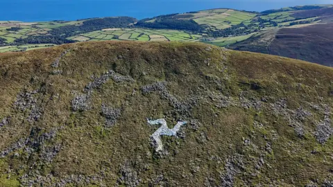 A fairly close view of the grassy hillside with a ridge that stretches from left to right, with a mass of green fields below in the distance and the sea beyond. Below the top of the ridge of North Barrule is a white Three Legs of Man symbol, clearly visible on a sunny day.