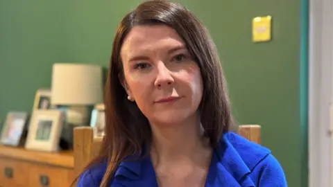 Sinead Lunny has shoulder length brown hair, wearing a blue blouse, looking down the camera. She is sitting on a brown chair with green wallpaper in the background and several framed photos on a dresser.