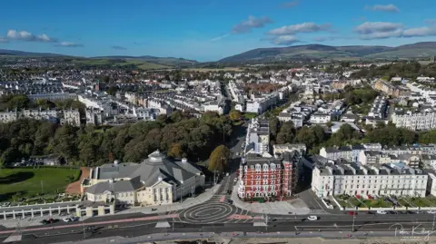 An arial view of Douglas, you can see a lot of buildings, the Villa Marina is at the front and you can see a next to it, there are hills in the background.