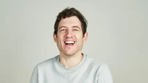 Elis James con cabello y rastrojo marrón corto, con un jersey gris, sonríe a la cámara