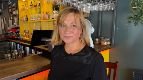 A woman with short blonde hair and a fringe who is wearing a black blouse. She is standing in front of the bar and looking at the camera.