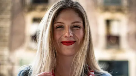 A young woman with long blonde hair and red lipstick smiles at the camera.