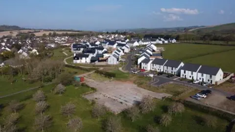 A drone view of the site. It is a large estate with Greenland surrounding the area. There is many white buildings with large grey roofs.