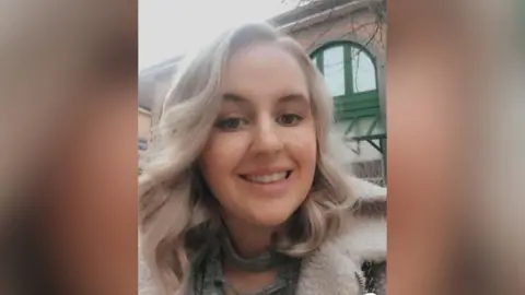 Abbigail Smith stands outside a brick building with large curved windows. She has light blonde hair down to her shoulders and is wearing a fur-lined jacket. She is smiling at the camera.
