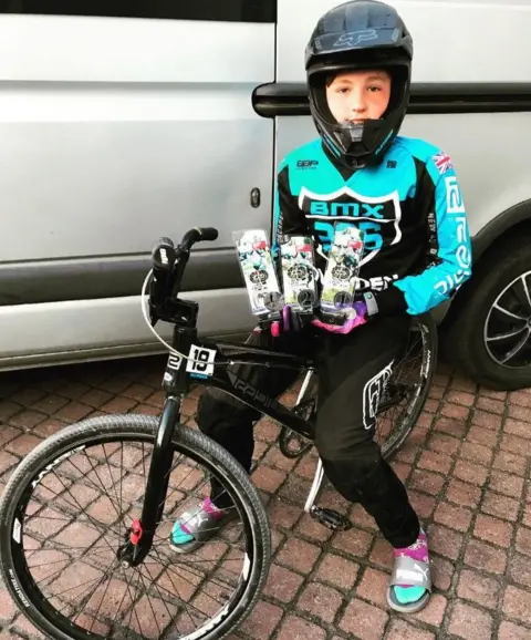 Supplied A boy in a teal and black BMX racing outfit and his helmet on his head. He is sat on his BMX bike with three awards in his hands. 