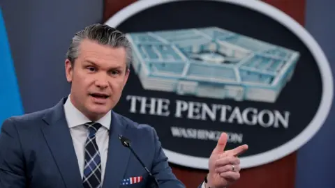 U.S. Secretary of Defense Pete Hegseth, in a dark suit, stands in front of The Pentagon sign