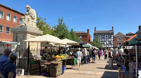 BBC Lichfield town centre, with market stalls set up and people milling around. There is a clear blue sky, and a number of buildings surround the area where the market is being held.