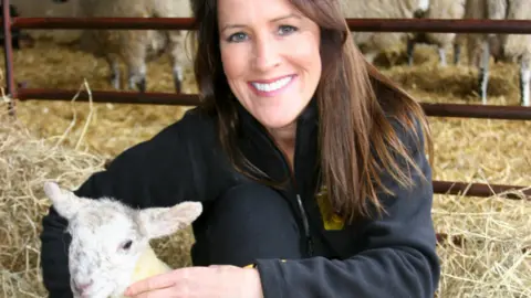 Alex Dunlop/BBC Stephanie Berkeley is smiling at the camera. She has long, brown hair and is wearing a dark blue top. She is holding a lamb.