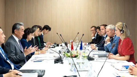 Getty Images Wang Yi and Kaja Kallas sitting at opposite finishs of a table, facing each other. They are surrounded by Chinese and European officials.