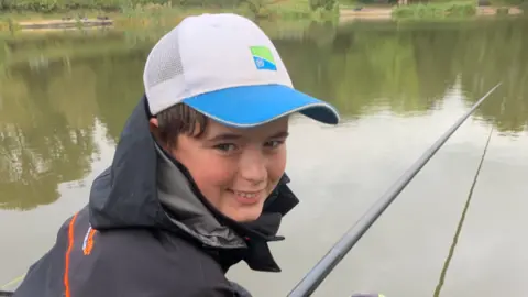 BBC Charlie smiles at the camera, wearing a white and blue cap and a black coat with his fish rod hanging over the lake in the background.