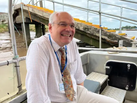 A man on board the King's Lynn ferry. He is wearing a white top with a red check pattern. His trousers are cream. He is smiling and wearing black glasses.