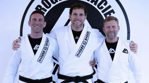 Clayton Chamberlain Three men wearing white gis standing shoulder to shoulder, in front of a club logo. All three have black belts.