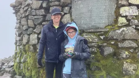 BBC Isabella and her mum standing in front of the trig point on the summit on an inclement day, both wearing rain coats with baseball caps on. 