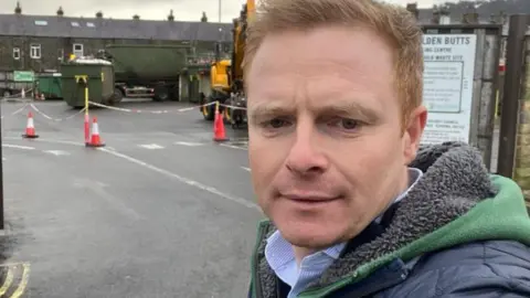 Robbie Moore A man with short ginger hair and a green hoodie, with a navy padded jacket over the top. He is taking a selfie outside a waste recycling centre in Ilkley. 