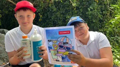 Ollie, 10, and Lewis, 9, smiling wearing uniform and sun hats. They are holding suncream and a sun safety colouring sheet. 