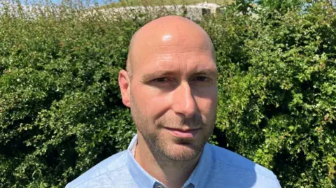 Neil Carlin is a bald man, he is wearing a blue shirt and standing in front of some bushes.