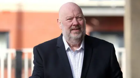 Former mayor of Liverpool Joe Anderson dressed in a suit and a white checked shirt. He has a grey beard and bald head. He is holding his hands together. 