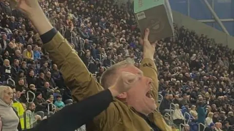 A man in a beige jacket holds a toaster in the air at a football ground