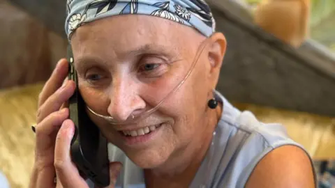 Lobular Moon Shot Dr Susan Michaelis, wearing a blue bandana and a light blue sleeveless top, speaks on a mobile phone, with a breathing tube attached to her nose.
