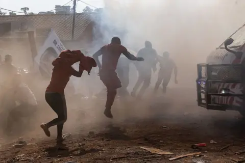 AFP via Getty Images A cloud of tear gas silhouettes a group of people as they try to run over dusty ground to avoid the smoke.