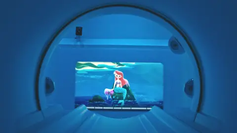 A view from inside an MRI scanner, looking through an archway with a cartoon image of a mermaid with red hair and a blue tail sitting on a rock in the sea at the end of the room. The room is lit in blue light.