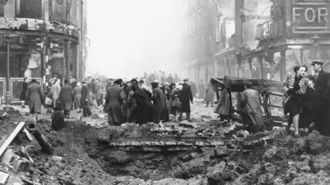 A black and white archive photo shows a huge hole surrounded and filled with rubble, in the middle of a road. There are two large buildings either side of the hole, lining the street. The buildings themselves are destroyed with only the external structures remaining. The remains of a sign saying FOR is on the end of one building. A crowd of people walking down the street and in the distance. A man and woman trying to walk around the edge of the hole.