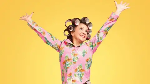 Andy Robinson A woman with brown hair in curlers is wearing a pink shirt with a floral pattern on it. She has her arms raised up in the air and is smiling. The background is yellow.