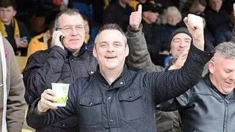 Family handout Simon Dobbin in the stand at a football stadium and holding a cup of tea. He has a fist in the air and is celebrating. He has short greying hair and is wearing a black coat in front of other football supporters.