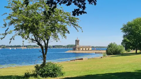 A church surrounded by a blue lake can been seen at Rutland water on summer day, with trees and grass on the right of the photo.