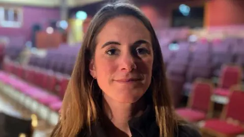 Katie Dyke has long brown hair and a black jacket. She is standing in an auditorium-type space with rows of seats.