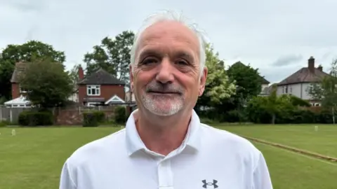 Charlie Stubbs/BBC There is a man with white hair, a grey stubbly goatee and a white under armour polo shirt. He is stood infront of croquet greens with a brown divider down the middle. There are three white croquet stumps, and a line of houses behind the fence.