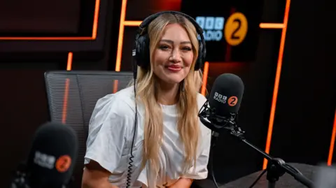 Hilary Duff in the Radio 2 studio. She has blonde hair and wears a white T-shirt