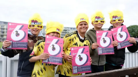 Getty Images Young Gloucestershire cricket fans, some of them wearing the team's T20 Vitality Blast kit, which is yellow with some faded black patches, hold up pink and white cards with the number 6 on them. The fans are at the County Ground in Bristol for the match with Somerset