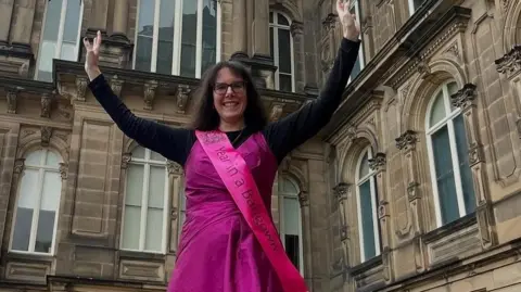 Supplied Jennifer Gane who has black glasses and long black hair, is wearing a pink ballgown over a black shirt. She is standing in front of the Bowes Museum building with her hands raised.