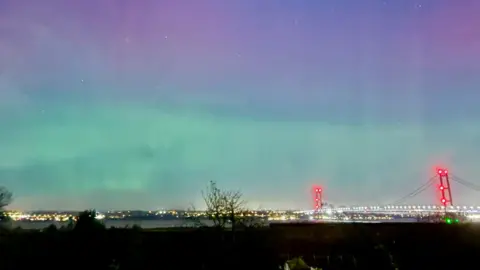 Humber Bridge lit up at night with red and white lights and the sky is purple and blue. There is a river and trees.
