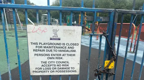 BBC/Claire Hamilton A sign on blue railings states that the playground has been closed for maintenance due to vandalism. There is a large metal chain around the gates, holding them closed. 