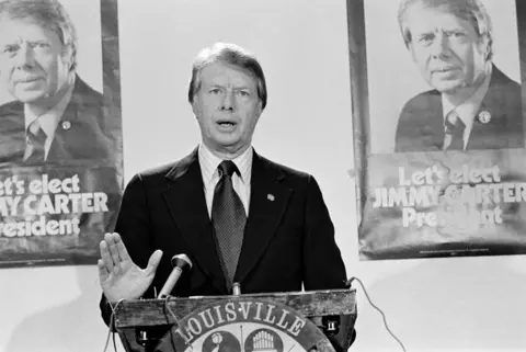 Getty Images Pastaj Guvernatori i Gjeorgjisë, Jimmy Carter qëndron përpara posterave që thonë "Le të zgjedhim JIMMY CARTER President" dhe i drejtohet Konventës për Çështjet Demokratike Kombëtare, 23 nëntor 1975