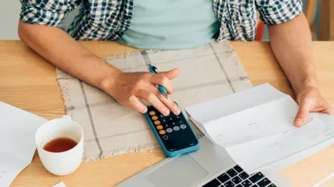 Stock image of a man sitting at a wooden table using his mobile phone to calculate figures in a document. There is also a laptop on the table and a cup of coffee to the left of the image.