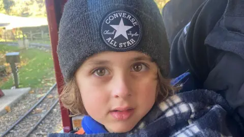 Reeve sits in a train carriage and smiles at the camera. He wears a grey beanie hat and chequered fluffy coat.