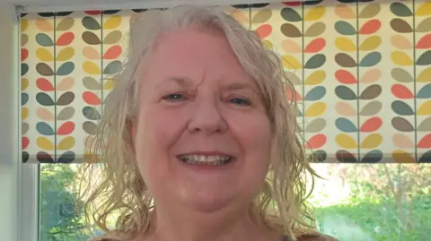 Kerry Oddy A woman with white and brown hair is smiling at the camera. She is near a window inside and a blind containing various colours is partially pulled down.