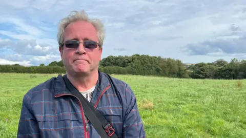 John Allinson standing in the field where he collapsed. He is wearing a checked-jacket and sunglasses, and has short grey hair.