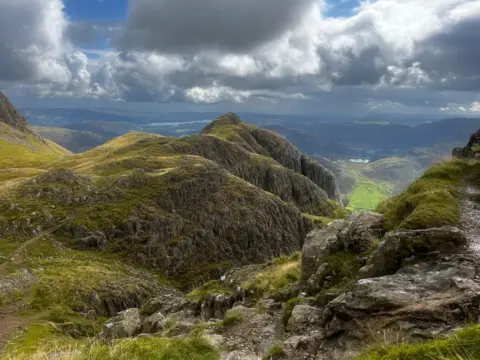 An image taken from the top of Langdale Fell, looking down towards the valley. The area is rocky but covered in lush green grass. The sky is heavy with grey clouds and a bit of haze looking towards a tarn and faraway fells.