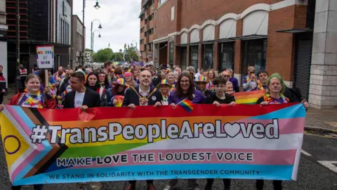 Wolverhampton Today People walking towards the camera through a city centre street as part of a parade. Many are wearing rainbow colours and purple shirts. One man at the front is wearing a gold mayoral chain. Six people at the front are holding a large multi-coloured banner which says "#TransPeopleAreLoved, make love the loudest voice, we stand in solidarity with the transgender community".